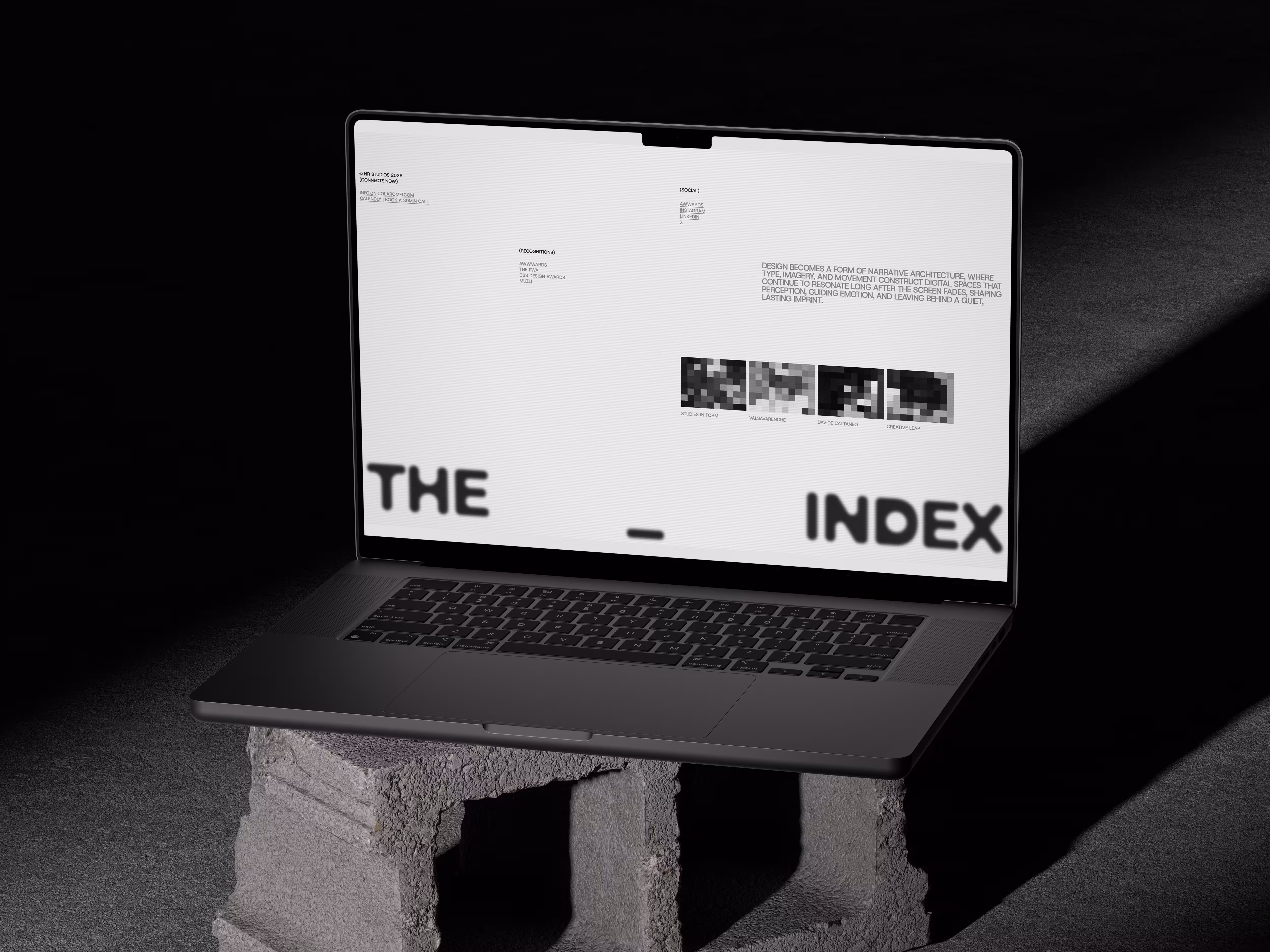 Laptop showing a webpage titled 'THE INDEX' with design text and pixelated images, on a cinder block.